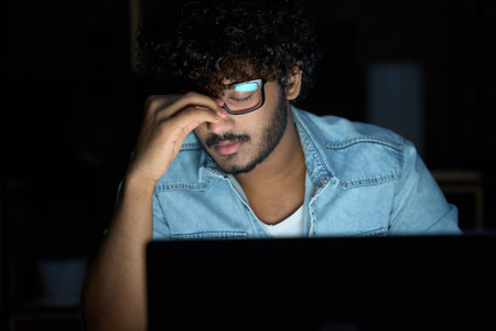 Tired sleepy young indian man wearing glasses using computer working late.