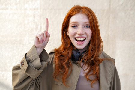Happy teen redhead girl standing on urban wall background pointing up.の写真素材