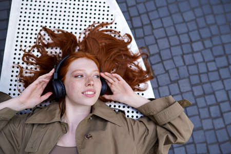 Teen redhead girl wears headphone listening music outdoor looking at copy space.