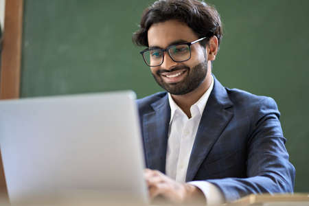 Smiling indian young business man executive wearing suit working on laptop.