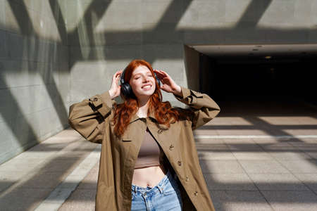Happy teen redhead girl wearing headphones listening music near urban walls.