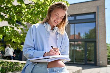 Happy girl university student writing in notebook studying sitting outdoor.