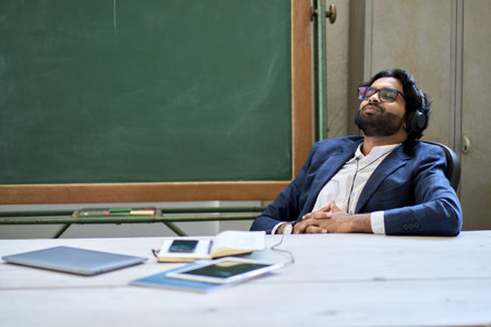 Young relaxed indian business man wearing headphones sitting listening to music.