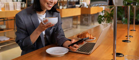 Young Asian business woman wearing suit drinking coffee using phone in cafe.