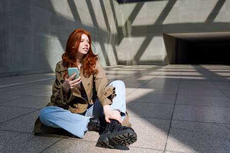 Redhead cool teen girl using phone sitting on urban city walls background.