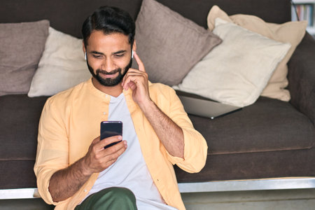 Indian man sitting at home on sofa using mobile phone watching videos.の写真素材