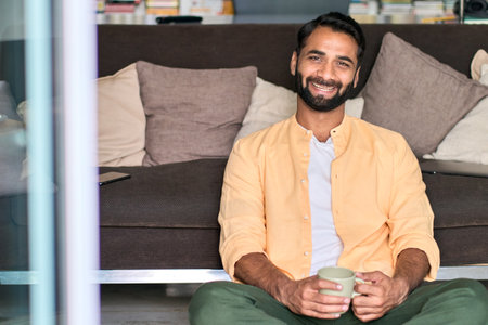 Happy indian man looking at camera drinking coffee sitting on sofa at home.の写真素材