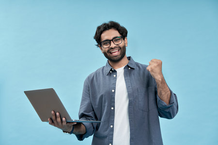 Happy young arab man celebrating win holding laptop isolated on blue.の写真素材