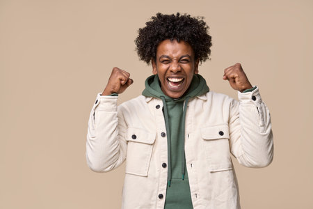 Happy excited African teen student winner boy celebrating win isolated on beige.