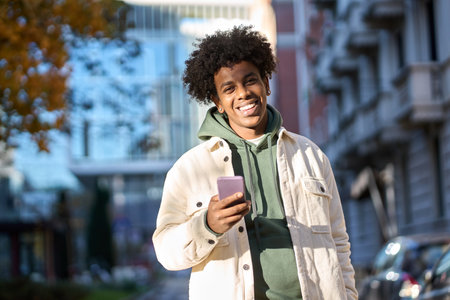 Happy young African American guy using phone standing at city street.
