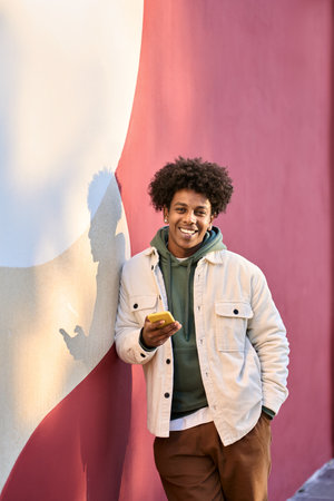 Positive African American hipster guy standing at red wall using mobile phone.の写真素材