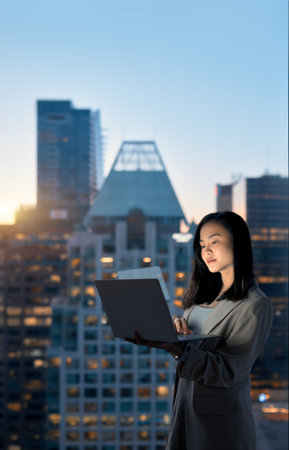 Young Asian business woman holding laptop standing at night office window.の写真素材