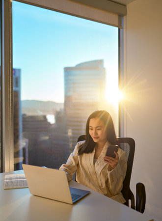 Young Asian business woman working using phone laptop in sunny office, vertical.の写真素材