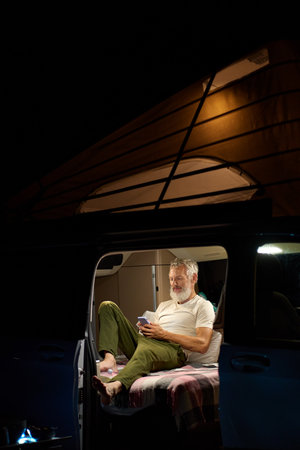 Older man traveler sitting in camper van using mobile in camping at night.の写真素材