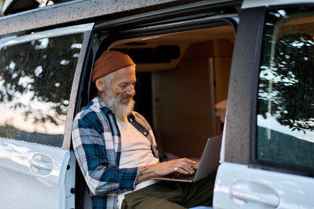 Happy older man traveler sitting in camper van using laptop in camping.の写真素材