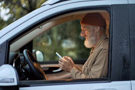 Happy older man traveler sitting in camper van using mobile in camping.