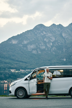 Mature man traveler standing near camper van using mobile in nature park.
