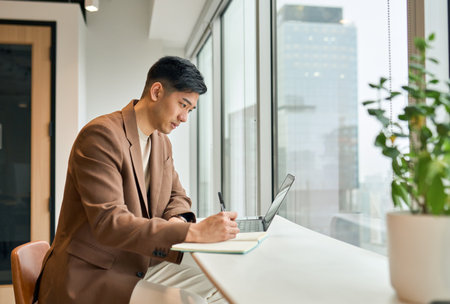 Young busy Asian business man working on laptop in office writing notes.