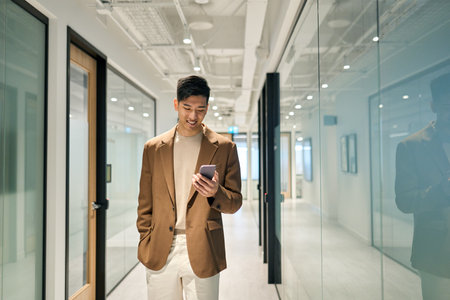 Young happy Asian business man standing in office hallway using phone.