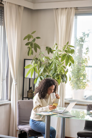 Young pretty woman sitting at cozy home holding phone using laptop.