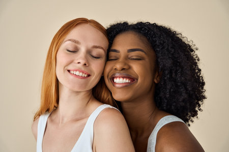Two smiling diverse cute young girls with eyes closed on beige background.の写真素材