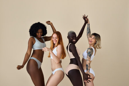 Happy multiracial girls wearing underwear dancing together on beige background.の写真素材