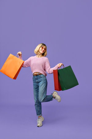Happy gen z blonde young woman holding shopping bags isolated on purple.の写真素材