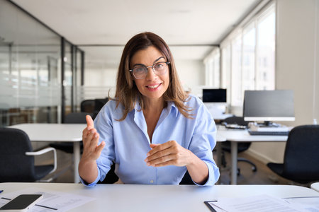 Middle aged woman executive talking to web cam sitting at work desk.