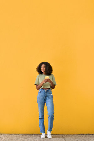Young happy African gen z girl standing at yellow wall using mobile phone.の写真素材