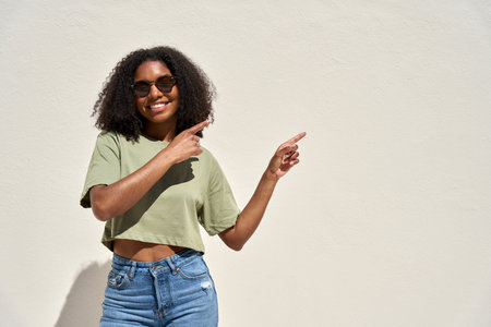 Smiling Afro American girl in sunglasses pointing aside standing at white wall.の写真素材