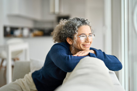 Happy mature woman sits on sofa in living room at home looking through window.の写真素材