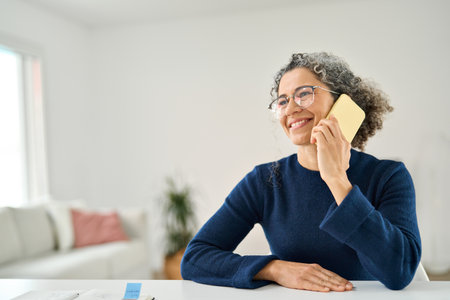 Happy middle aged woman making phone call sitting at table in living room.の写真素材