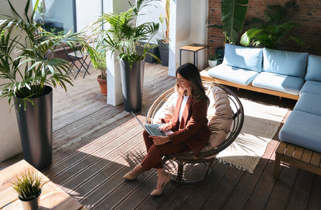 Busy mature business woman using laptop sitting in chair in sunny green office.の写真素材