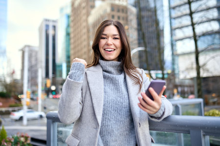 Happy young woman winner using phone winning online standing on street.