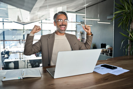 Excited mature business man celebrating goals achievement at work in office.