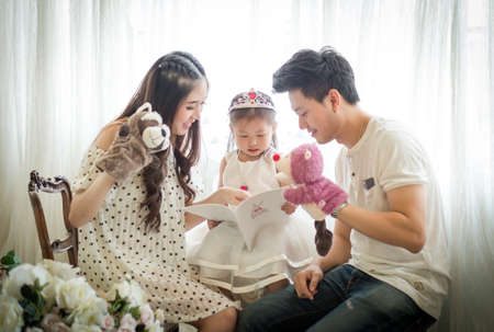 family, child and home concept - smiling parents and little girl with Storytelling at home.Backlit shotの写真素材
