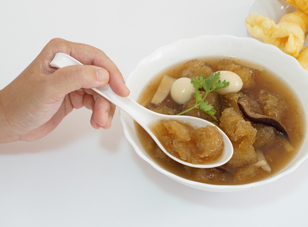 An Asian-forties-lady's hand is using a spoon to scoop Chinese style soup; or braised fish maw in red gravy with eggs, mushroom, and bamboo shoot in white bowl. White background.の写真素材