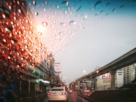 Blur street traffic in twilight after rain (Thailand); wet car glass (clear with blue color on top) from raindrops and dried part from windshield wiper. Skytrain rail on the right. Top copy-space.の写真素材