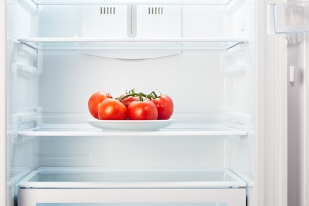 Branch of red tomatoes on white plate in open empty refrigerator. Weight loss diet concept.の写真素材