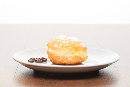 Fresh doughnut with icing sugar and coffee beans on white ceramic plate on bright light brown wooden table backgroundの写真素材