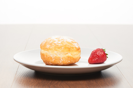 Fresh doughnut with icing sugar and strawberry on white ceramic plate on bright light brown wooden table backgroundの写真素材