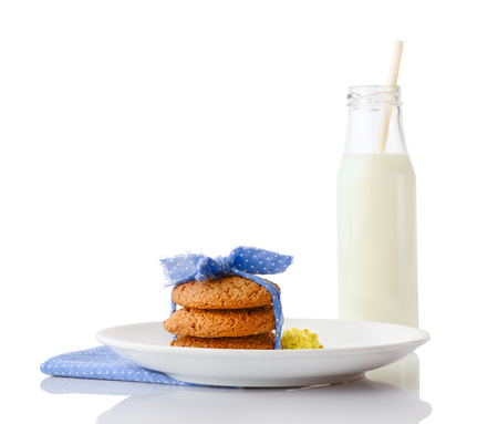 Stack of three homemade oatmeal cookies tied with blue ribbon in small white polka dots and tiny yellow flowers on white plate on napkin and bottle of milk with straw, isolated on white backgroundの写真素材