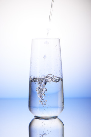 Water pouring into half-filled drinking glass with reflection on white and blue backgroundの写真素材
