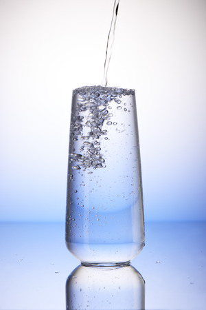 Water pouring into full drinking glass with reflection on white and blue backgroundの写真素材