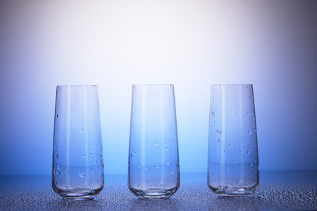 Three empty drinking glasses with reflection in drops of water on white and blue backgroundの写真素材