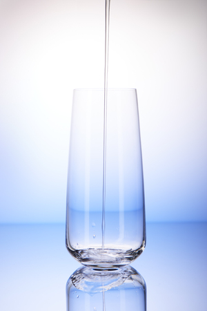 Water pouring into empty drinking glass with reflection on white and blue backgroundの写真素材