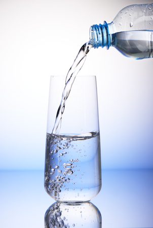 Water pouring from plastic bottle into half-filled drinking glass with reflection on white and blue backgroundの写真素材