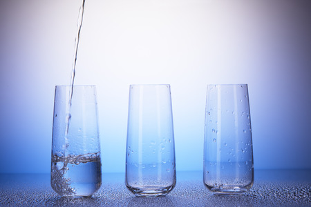 One-third full and two empty drinking glasses with reflection in drops of water on white and blue background. Water pouring into first one.の写真素材