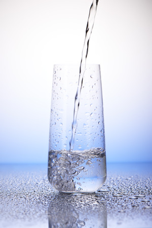 Water pouring into one-third full drinking glass with reflection in drops of water on white and blue backgroundの写真素材