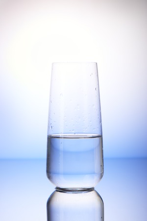 Half-filled drinking glass with reflection in drops of water on white and blue backgroundの写真素材